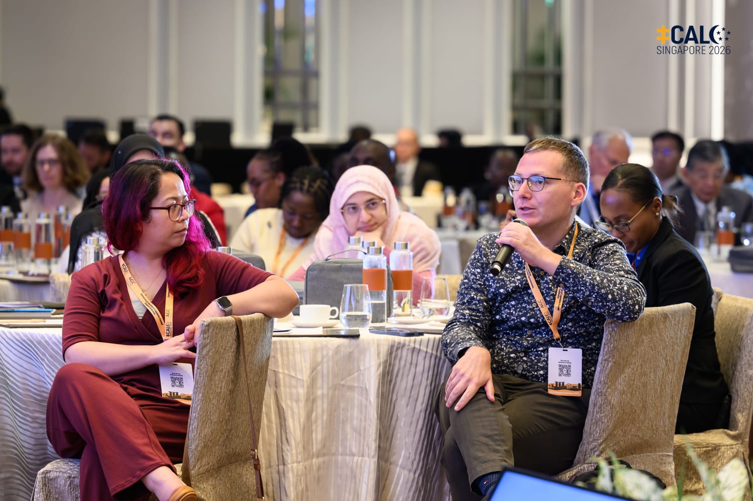 Two conference attendees seated at a round table engage in discussion, with a man holding a microphone, at the CALC Singapore 2026 conference. A diverse group of delegates is visible in the background.